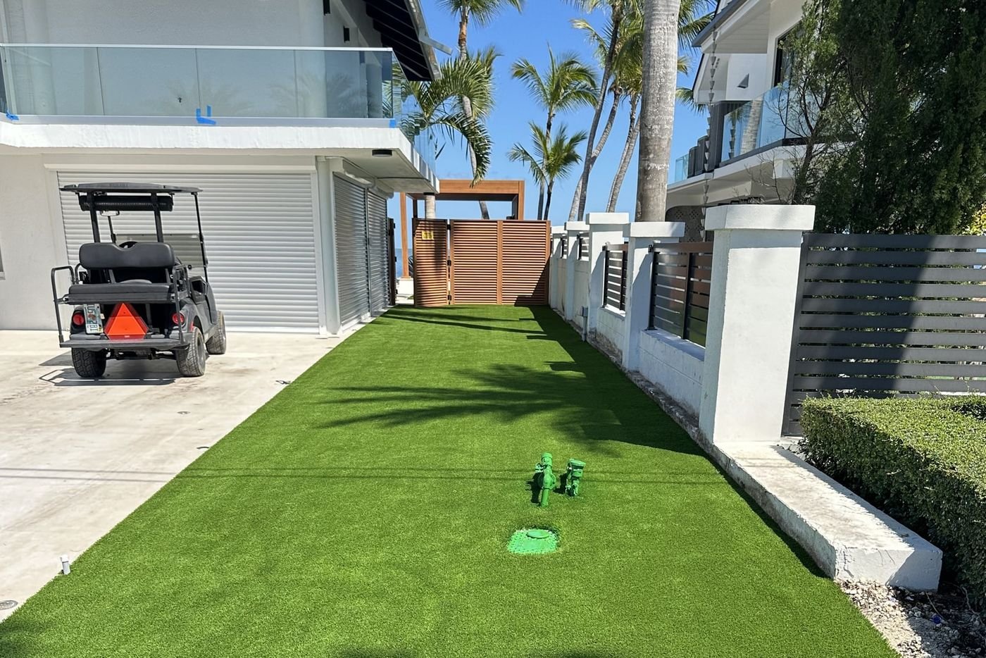 Franja de grama artificial junto a la entrada de coches de una casa moderna, con un carrito de golf y palmeras al fondo.