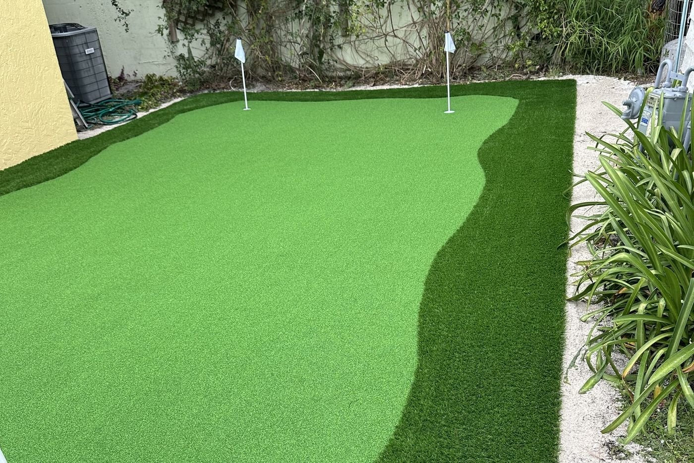 Putting green de grama artificial con dos banderas, delante de un muro cubierto de enredadera.