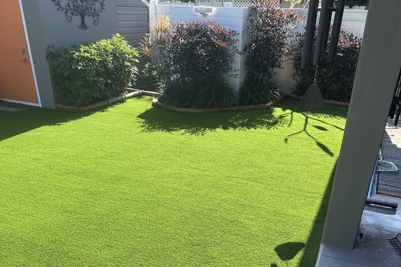 Patio con grama artificial, una caseta de jardín gris y arriates curvos con arbustos.