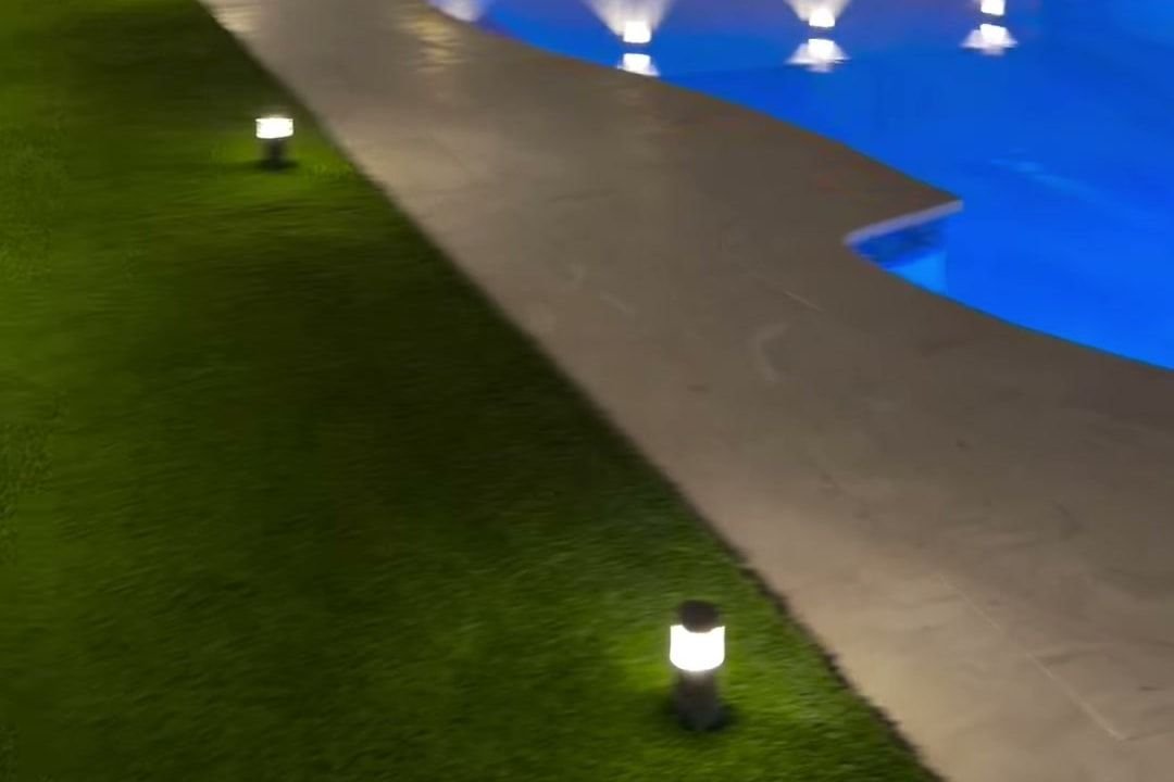 Patio con piscina iluminada de noche y grama artificial junto al pavimento.