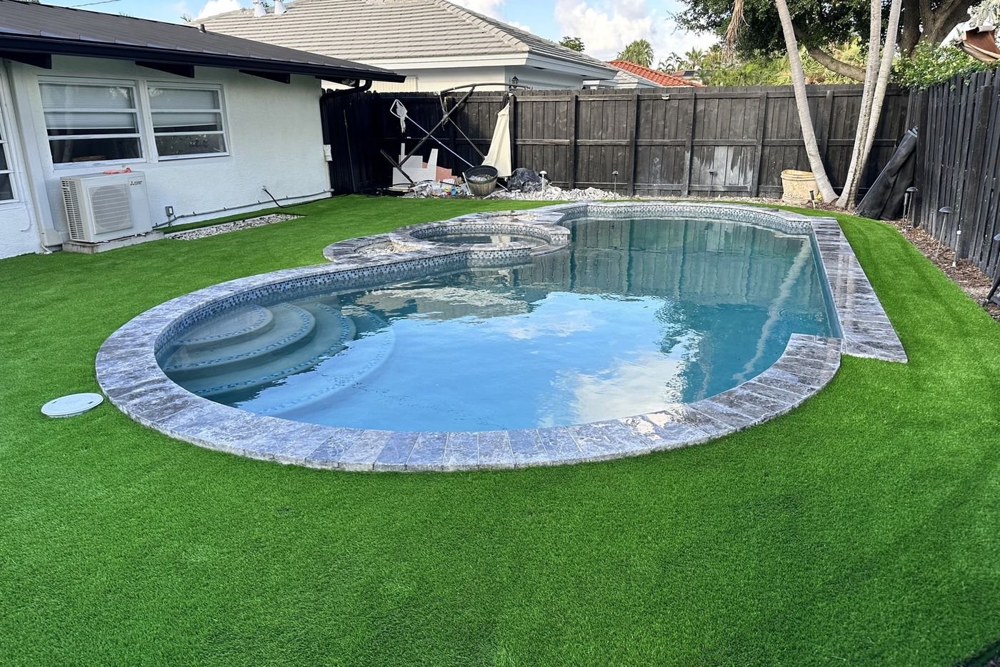 Patio con grama artificial rodeando una piscina curva con borde de piedra, junto a una casa de una planta.